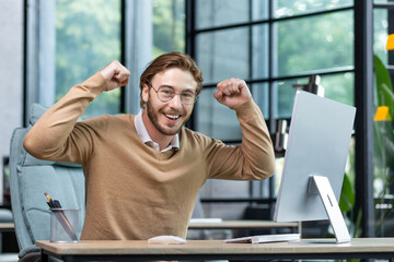 A young businessman, student, freelancer, programmer sits in the office at the table and celebrates success, victory. Joyfully raises his hands up and shows strength, looks into the camera, smiles.