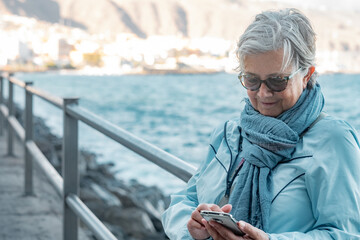 Senior woman outdoors on a winter day relax near the beach with her back to the ocean texting a...