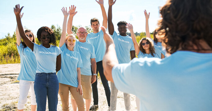 Volunteer leader planning activity, instructing volunteers on garbage, trash, rubbish collection. Multi-ethnic diverse of different ages raising hands and smiling. Biosphere, ecology, global problem.