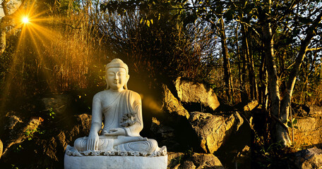 White meditating Buddha statue sits on the rock pile among forest trees in the afternoon time at...