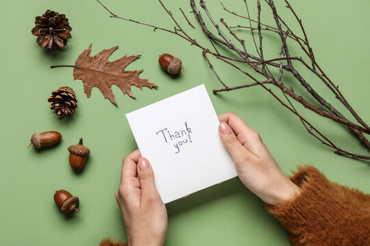 Woman holding paper sheet with text THANK YOU, acorns, fir cones and tree branches on green background