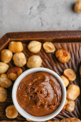 Wooden board with bowl of tasty hazelnut butter and nuts on light background, closeup