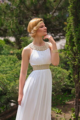 Fototapeta na wymiar blonde beautiful natural girl with blonde hair in a white dress in nature, on the street, in the garden by a tree, with makeup and hairstyle, greek woman in a white ethnic historical dress, retro