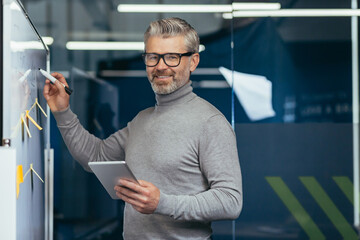 Portrait of happy and smiling businessman near white board inside office, senior man smiling and looking at camera holding tablet computer writing strategy plan on board with papers.