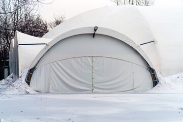 A large prefabricated hangar made of metal structures and synthetic film is covered with snow