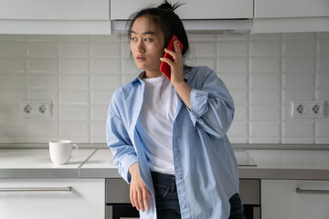 Tired pensive Asian woman standing in house trying to get through to boyfriend. Perplexed Korean girl putting mobile phone to ear waiting for answer from repairman after kitchen equipment breaks down
