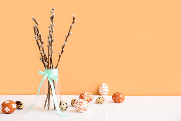 Glass vase with pussy willow branches, turquoise ribbon and Easter eggs on white table near orange wall