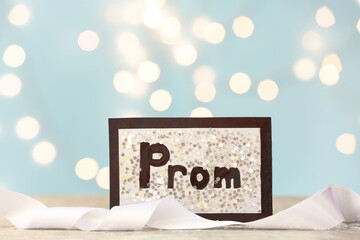 Card with word PROM and ribbon on table against blurred lights