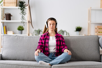 Young pretty woman meditating on sofa while listening to relaxing music on headphones at home