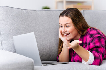 Fototapeta premium Pretty cheerful young woman having video call on laptop while lying down on sofa.