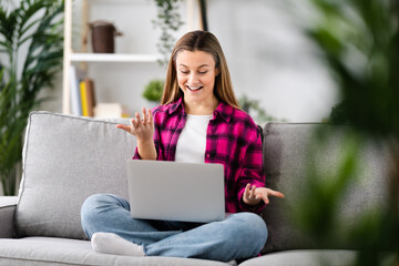 Pretty young woman having video call on laptop while sitting on sofa in the living room at home. Live streaming and homeschooling concept