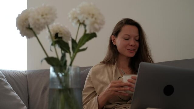 Young woman in bathrobe is sitting on sofa drinking coffee and making video call using a laptop in the foreground of bouquet of fresh flowers