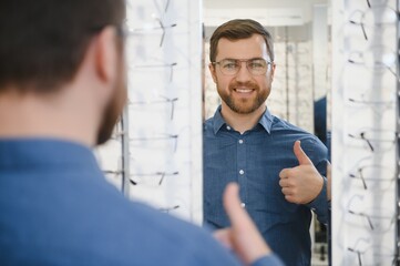 Satisfied Customer. View of happy young male client wearing new glasses, standing near rack and showcase with eyewear. Smiling man trying on spectacles