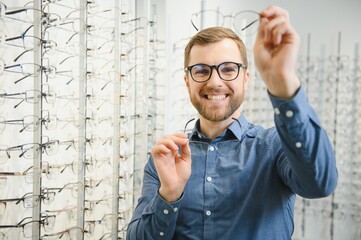 Satisfied Customer. View of happy young male client wearing new glasses, standing near rack and showcase with eyewear. Smiling man trying on spectacles