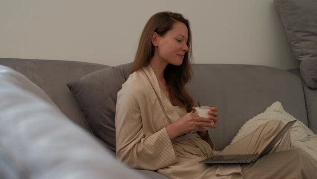 Side view of young woman in bathrobe is sitting on sofa at home drinking coffee and making video call using a laptop