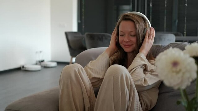 Young cheerful woman enjoying listening to music in headphones while sitting on sofa at home