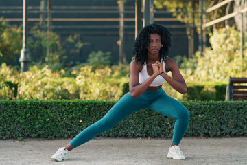 mujer de color negra con ropa deportiva, preparándose para hacer ejercicios, sentadillas, estirando, flexiones, corriendo en un parque con luz natural.	 - obrazy, fototapety, plakaty