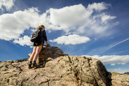 Female Hiker With Backpack Stands On A Rock Looking Into The Distance Under A Blue Cloudy Sky.