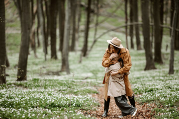 Front view of a happy stylish woman hugging her daughter in forest. Little girl in trendy clothes hugging with mother while spending time in spring park