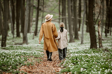 Back view of mother and daughter in trendy outfits walking in forest. Woman in beige trench and hat holding hands of little girl with bow on hair, during a stroll in woods with blooming flowers