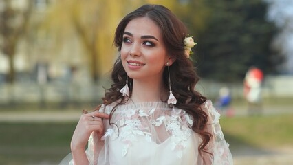 The bride in a white dress shows beautiful makeup on her face in the street.