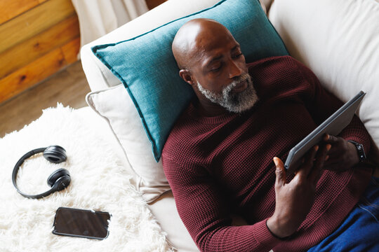 Bald african american senior man using tablet while lying on sofa in log cabin