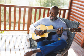 Bald african american senior man singing and playing guitar while sitting on chair in balcony