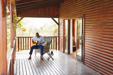 African american senior man playing guitar while sitting on chair with coffee on table in balcony
