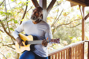 Bald african american senior man playing guitar while sitting on railing in balcony at log cabin