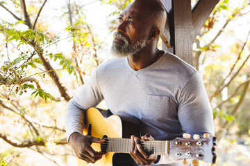 African american bald senior man playing guitar while sitting by column in balcony at log cabin