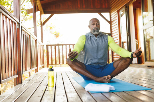 Bald african american senior man meditating on mat in balcony at log cabin
