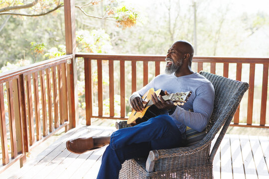 Bald african american senior man playing guitar while sitting on chair in balcony at log cabin
