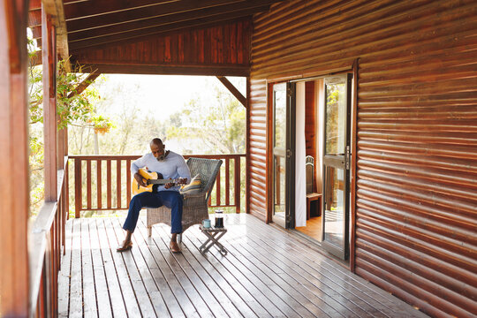 African american senior man playing guitar while sitting on chair with coffee on table in balcony