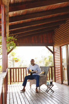 African american senior man playing guitar while sitting on chair with coffee on table in log cabin