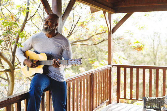 Smiling african american senior man playing guitar while sitting on railing in balcony at log cabin