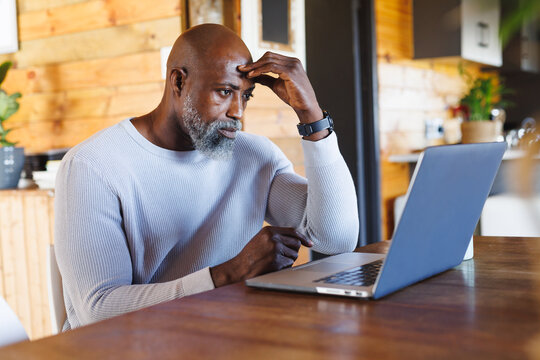 Stressed Bald African American Senior Man With Head In Hand Looking At Laptop In Log Cabin