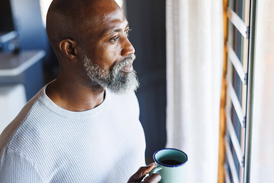 Thoughtful african american bald senior man with coffee mug looking through window at log cabin
