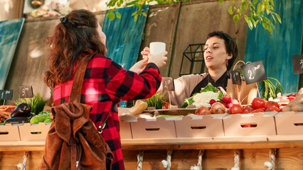 Stall holder drinking cup of hot coffee with regular customer, feeling happy with organic eco produce. Business owner sharing drink with client, selling homegrown products. Handheld shot.