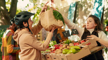 Farmers market vendor giving healthy food order to woman working as delivery courier, farmer helping clients. Young adult on bicycle delivering frutis and vegetables. Handheld shot.