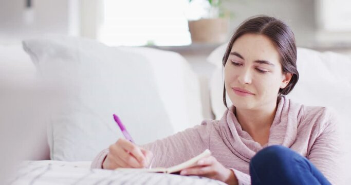 Happy caucasian woman sitting on floor and taking notes on couch