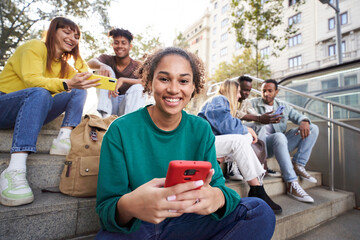 Smiling girl looking at camera holding her smart phone sitting on city street outdoors with...