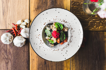 Top-down view of black tagliolini pasta with shrimp and sun-dried tomatoes served on white plate on wooden table decorated with garlic and peppers. Italian cuisine. Horizontal indoor shot. High