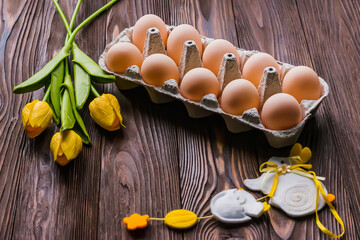 Cardboard tray with Easter eggs on a wooden table next to a bouquet of yellow tulips and Easter decor