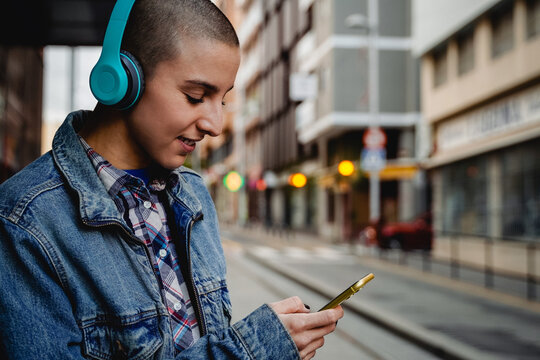 Bald young woman listen playlist music while waiting at bus station outdoor - Focus on face