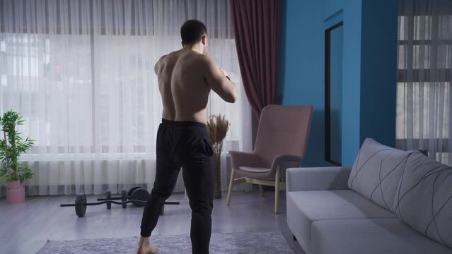 Young athlete doing kickboxing against the air at home with his back turned.
Muscular sportsman shadowboxing at home with his back turned to the camera, kicking and punching.
