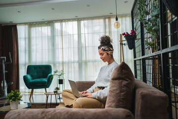 Obraz premium A young girl studying or doing business from her home while sitting on sofa and working on her laptop and drinking coffee during the day 