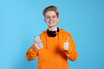 Teenage boy with smartphone and headphones showing thumb up on light blue background