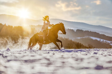 Fototapeta na wymiar An equestrian woman galloping on her horse through snow in front of a winter mountain landscape during sundown outdoors