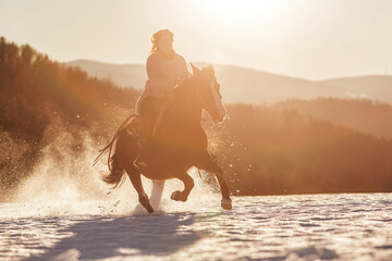 An equestrian woman galloping on her horse through snow in front of a winter mountain landscape...