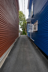 An alleyway or passageway between two tall wooden buildings. One is red and the other is bright blue. The exterior walls are made of wooden clapboard. There are trees at the end of the alley. 